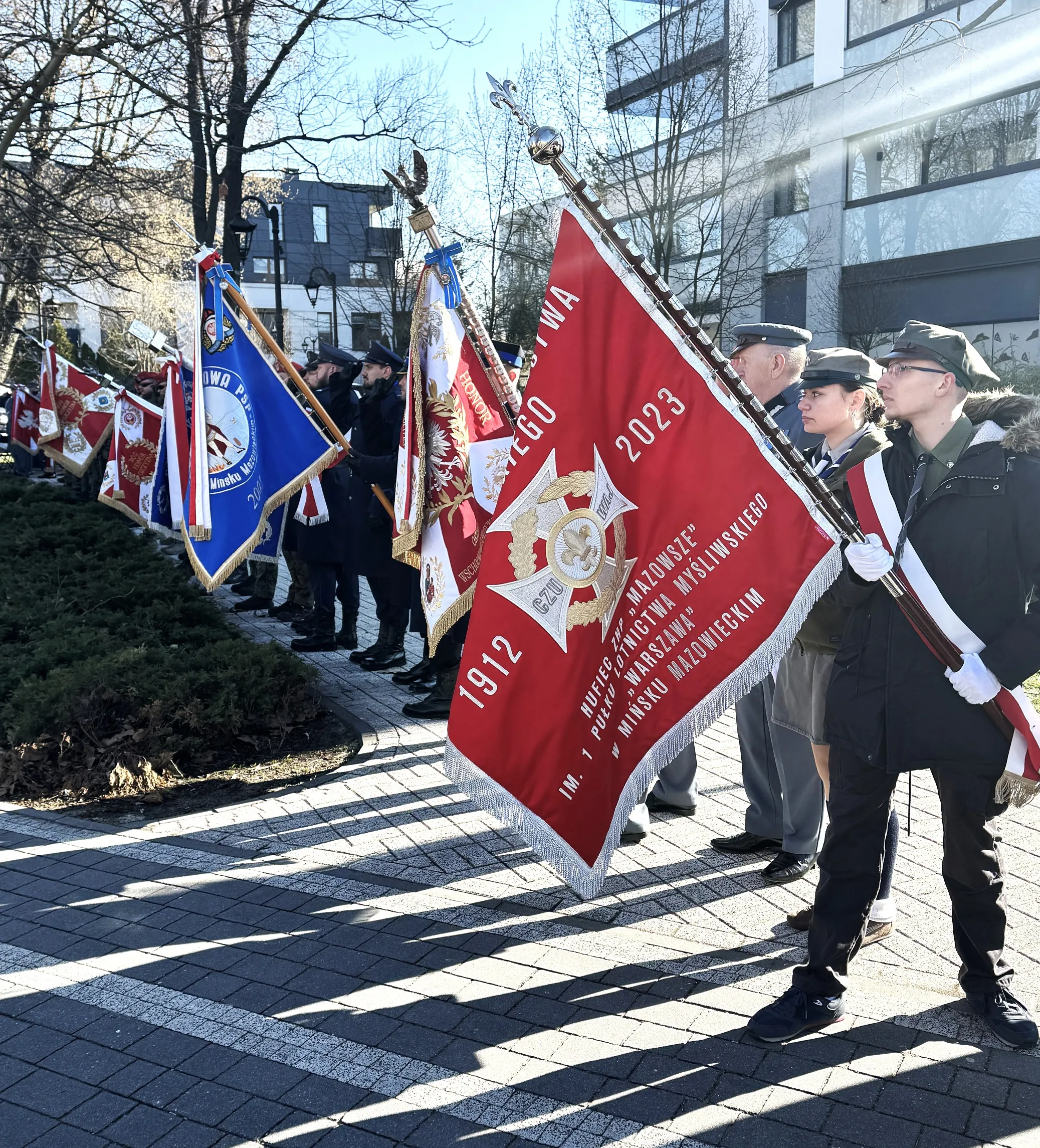 Grupa osób w mundurach stoi w rzędzie na zewnątrz w dzień. Osoby trzymają duże, zdobione proporce w czerwieni i błękicie. W tle nowoczesne budynki, nagie drzewa i chodnik z kamienną nawierzchnią.