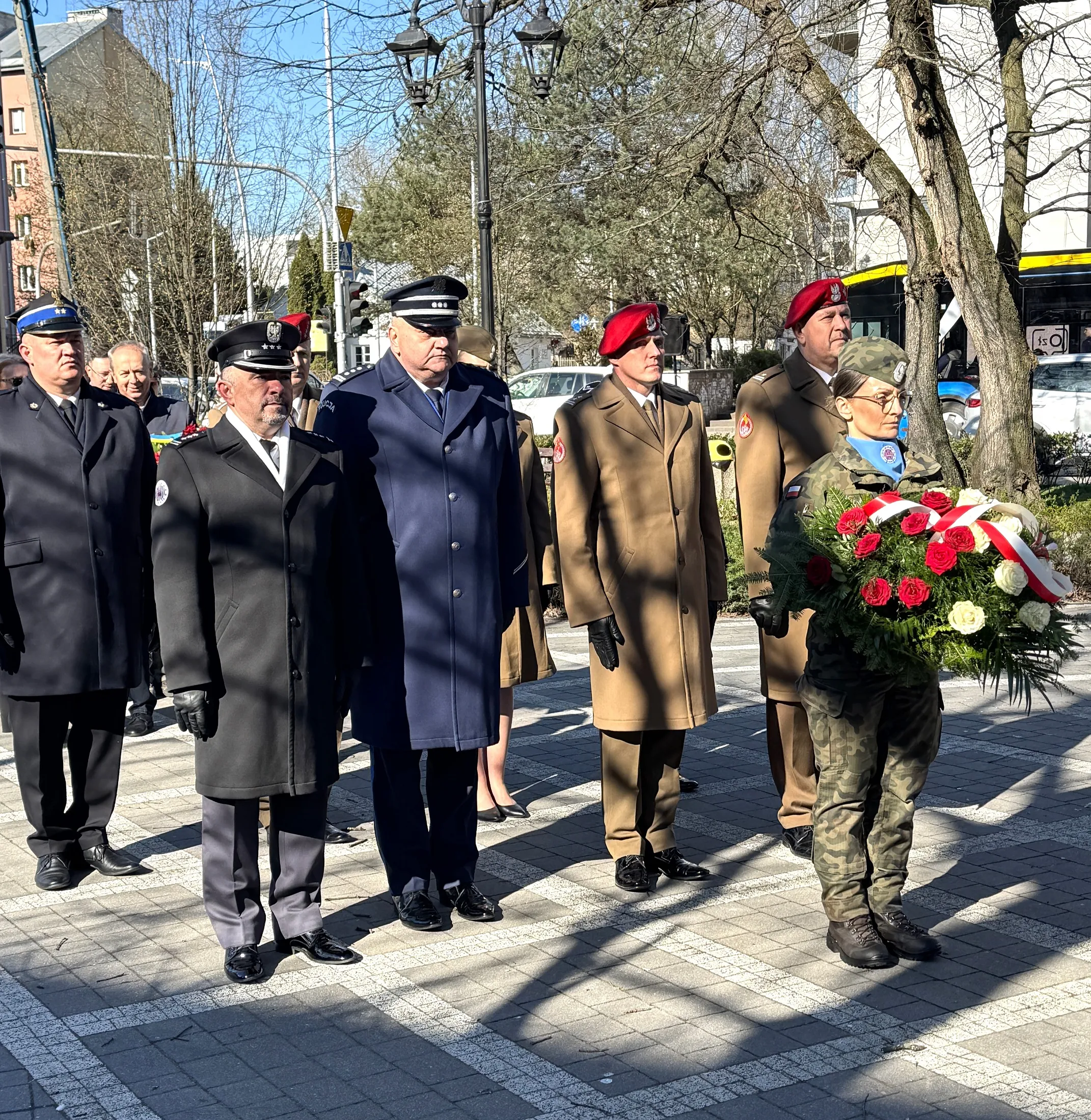 Sześć umundurowanych osób stoi w szeregu na zewnątrz w dzień na wybrukowanym placu. Po prawej kobieta w mundurze kamuflażowym niesie wieniec z czerwono-białych róż. Po lewej mężczyźni w granatowych i brązowych płaszczach oraz w czapkach i czerwonych beretach. W tle drzewa, latarnie i przejeżdżający autobus.