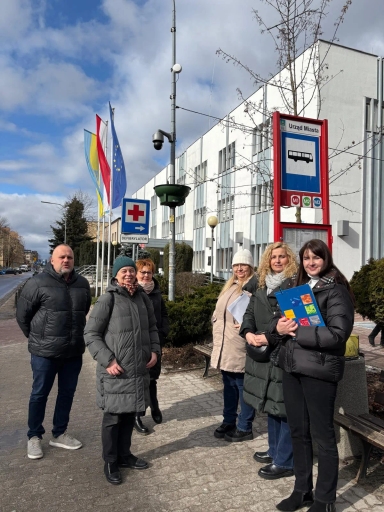 Sześć dorosłych osób stoi na chodniku przed białym budynkiem urzędu miasta. Osoby noszą zimowe kurtki i stoją przy przystanku autobusowym z niebieskim znakiem. Po lewej widać kilka pionowych flag, w tym flagę Unii Europejskiej. Na słupie widoczny jest znak z czerwonym krzyżem i napis o defibrylatorze. Niebo jest częściowo zachmurzone.
