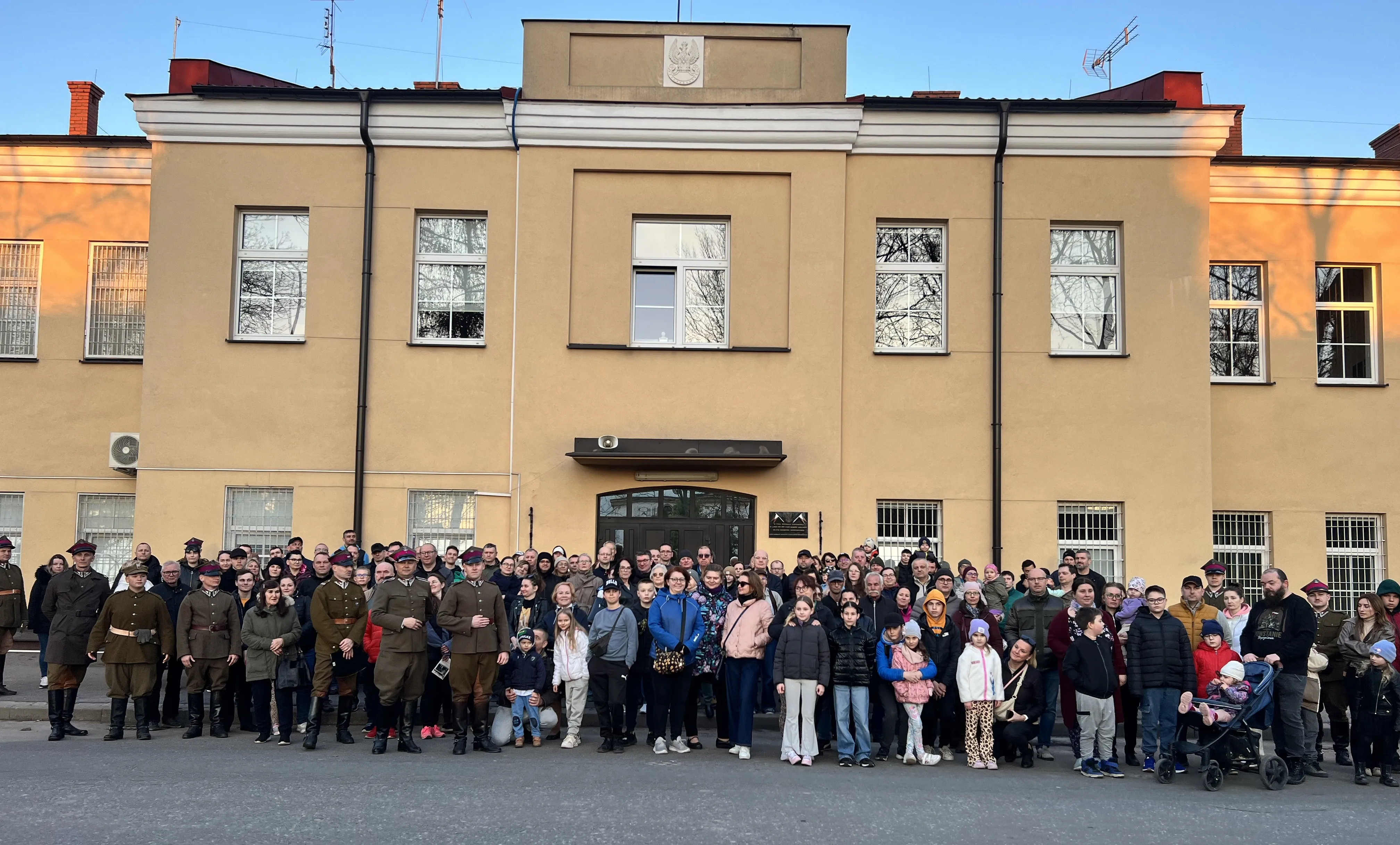 Duża grupa dorosłych i dzieci stoi przed dwukondygnacyjnym, beżowym budynkiem w ciągu dnia. Kilku mężczyzn w historycznych mundurach stoi z przodu po lewej stronie. W tłumie widać wózek dziecięcy, różne kurtki i okna budynku odbijające drzewa.