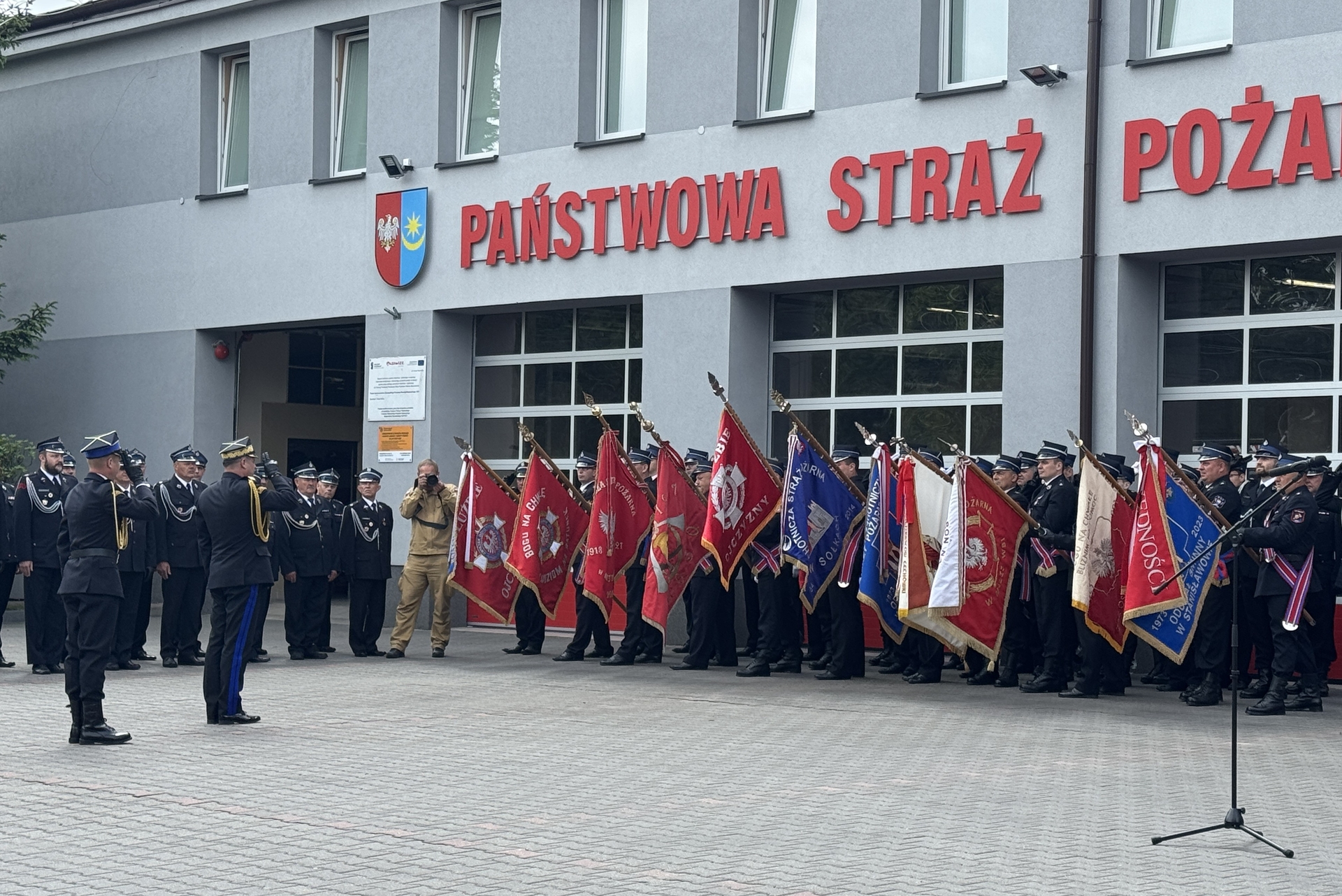 Grupa mundurowych stoi przed szarym budynkiem remizy. W szeregu kilkudziesięciu osób trzymają czerwone i niebieskie sztandary. Scena odbywa się na zewnątrz w ciągu dnia.