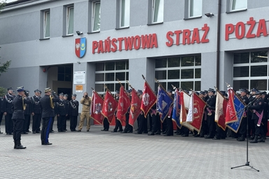 Grupa mundurowych stoi przed szarym budynkiem remizy. W szeregu kilkudziesięciu osób trzymają czerwone i niebieskie sztandary. Scena odbywa się na zewnątrz w ciągu dnia.