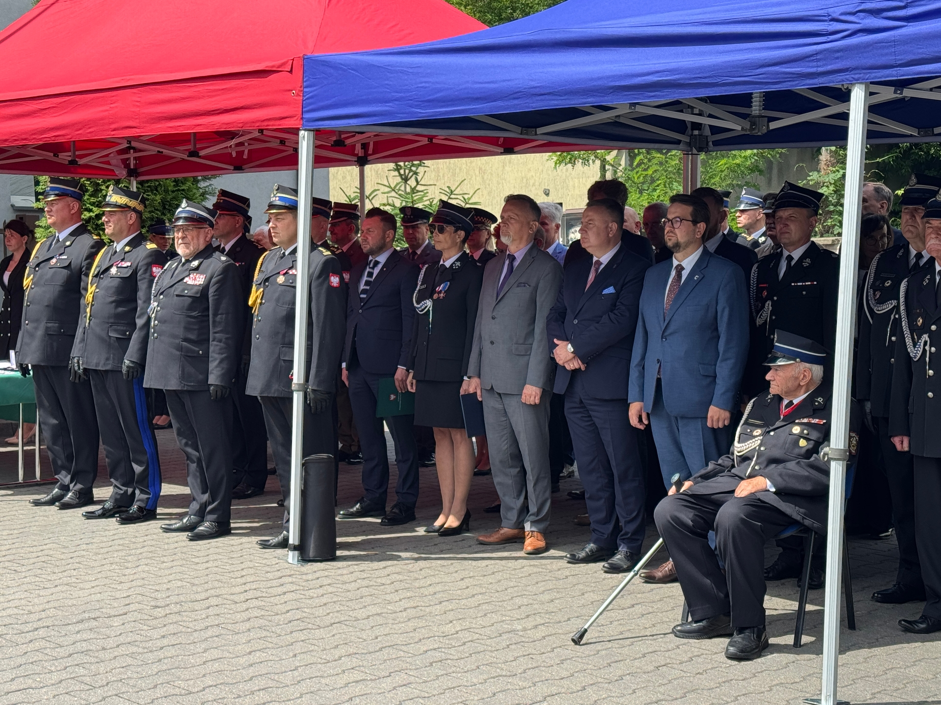 Grupa kilkunastu osób w mundurach i garniturach stoi w rzędzie pod połączonymi czerwonym i niebieskim namiotami. Funkcjonariusze noszą ciemne mundury i czapki, kilka osób ma medale. Na prawo siedzi starszy mężczyzna w mundurze z laską. W tle widoczne drzewa, budynek i brukowany plac.