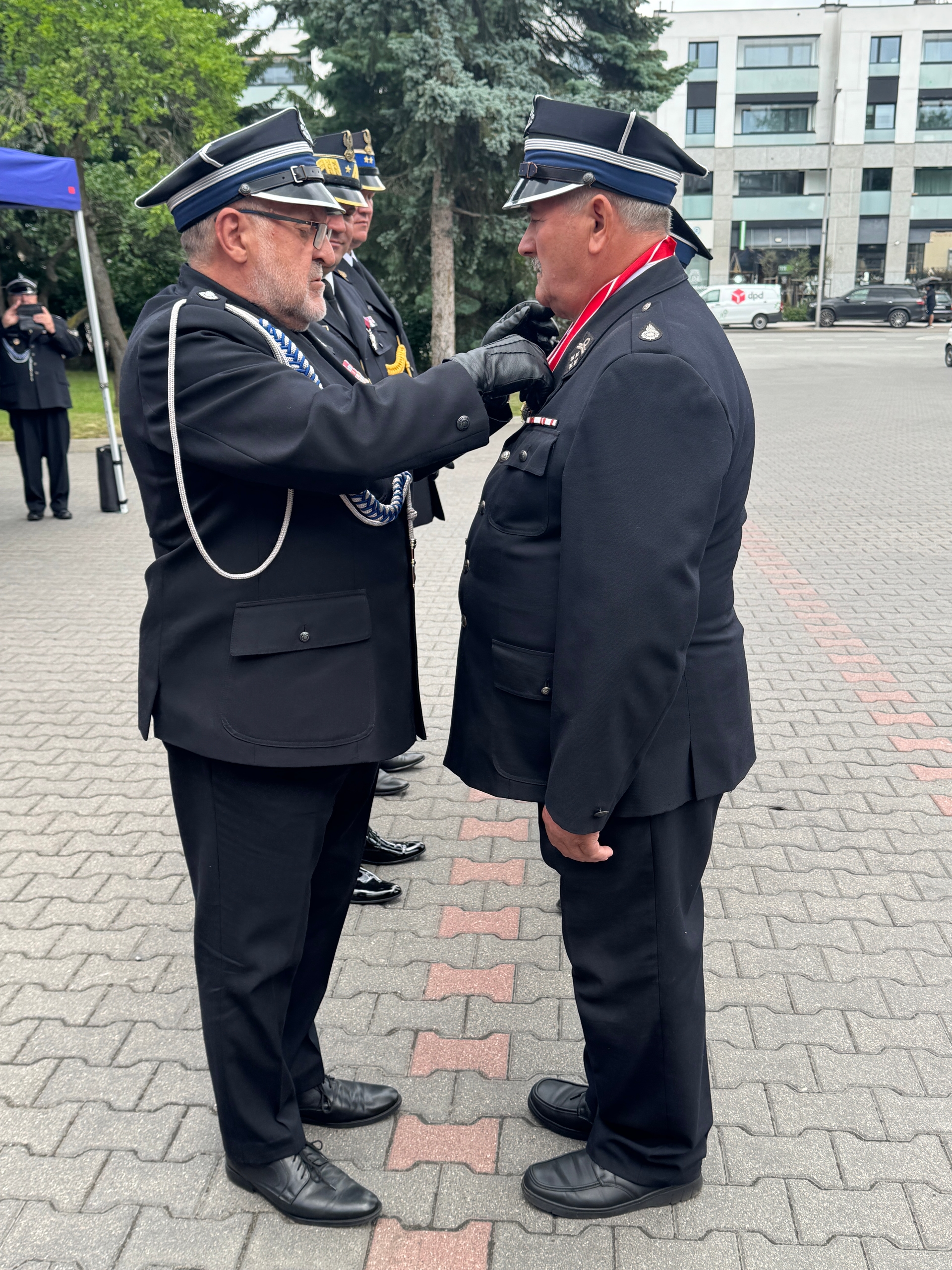 Dwaj umundurowani mężczyźni stoją twarzą w twarz na zewnętrznym placu. Starszy mężczyzna po lewej przypina medalik do munduru mężczyzny po prawej; w tle drzewa i budynek biurowy.