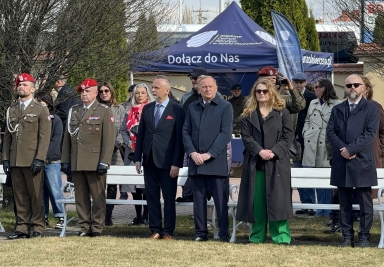 Sześcioro dorosłych stoi w szeregu na zewnątrz na trawniku przed białą ławką. Po lewej dwaj żołnierze w oliwkowych mundurach i czerwonych beretach. Po prawej kilku cywilów w płaszczach i garniturach. Za nimi niebieski namiot z białymi napisami i osoby w tle, w tym fotograf. 