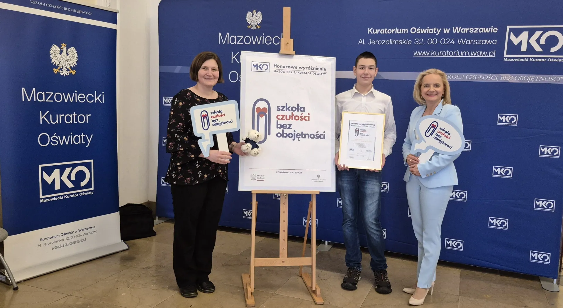 Trzy osoby stoją w pomieszczeniu z niebieskimi ściankami promocyjnymi Mazowieckiego Kuratorium Oświaty. Kobieta po lewej trzyma misiaczka i tabliczkę w kształcie dymka z napisem. Chłopiec w środku trzyma framed certyfikat. Kobieta po prawej uśmiecha się i trzyma podobną tabliczkę. Na środku stoi plansza na sztaludze z napisem "szkoła czułości bez obojętności".