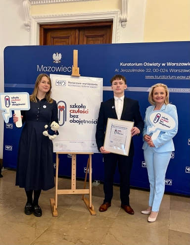 Trzy osoby stoją na tle niebieskiego banera z napisem „Mazowiecki Kuratorium Oświaty”. Kobieta po lewej stronie ma ciemną sukienkę, trzyma pluszowego misia i tabliczkę z logo i napisem „szkoła czułości bez obojętności”. Mężczyzna pośrodku w garniturze trzyma ramkę z wyróżnieniem. Kobieta po prawej stronie w jasnoniebieskim garniturze trzyma dużą tabliczkę z tym samym logo i napisem. Za nimi stoi plansza na sztaludze z duplikatem logo i napisem „Honorowe wyróżnienie Mazowieckiej Kurator Oświaty”.