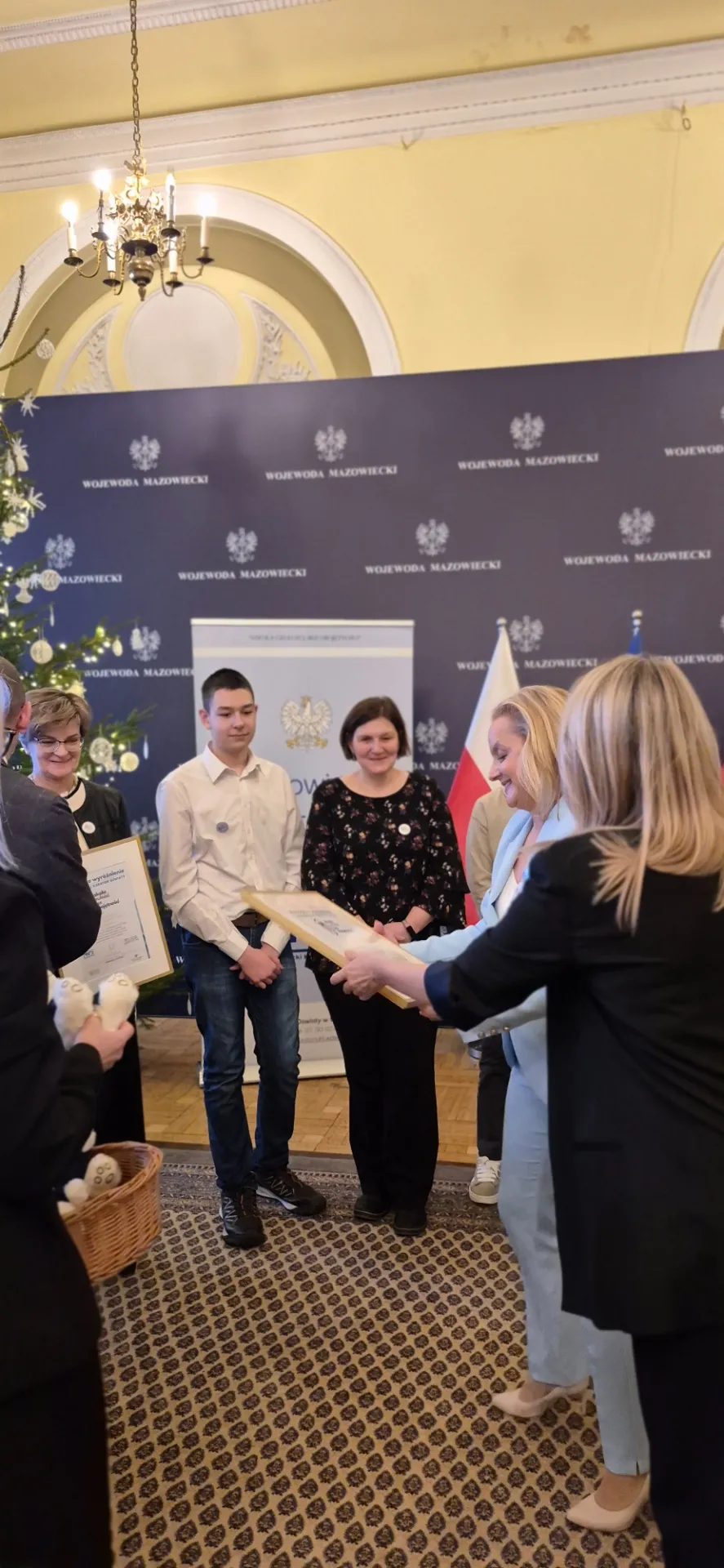 Grupa osób w jasnym pomieszczeniu z żyrandolem. Kilka osób stoi na wzór, w tym młody mężczyzna w białej koszuli i dziewczyna w niebieskim garniturze, która przekazuje ramę z dokumentem. W tle niebieska ściana z napisem "Wojewoda Mazowiecki" i polska flaga. Po lewej stronie dekorowana choinka i mężczyzna z koszykiem pluszaków.