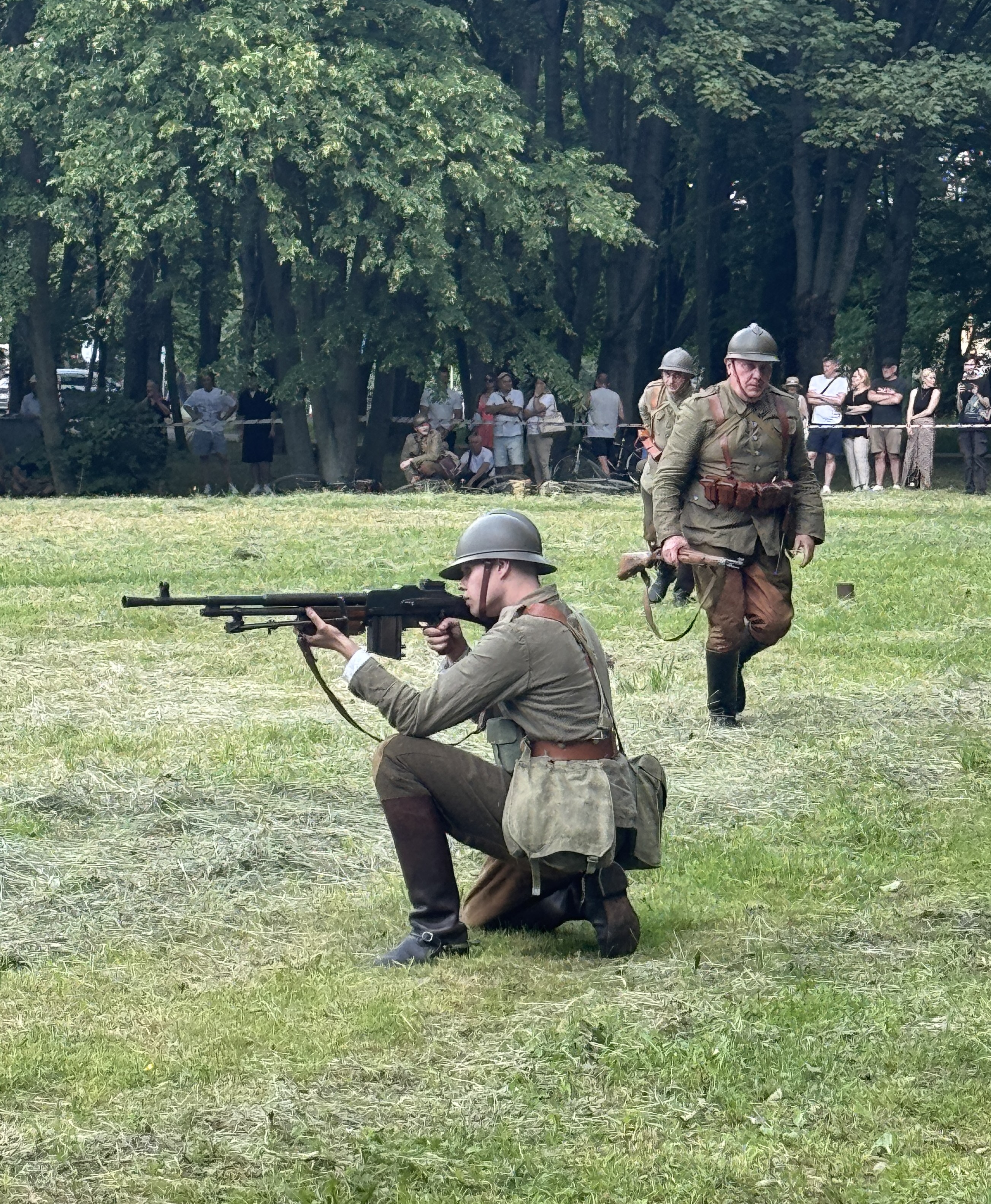 Dwaj żołnierze w historycznych mundurach i hełmach stoją na trawiastej polanie. Jeden klęczy i celuje ciężkim karabinem maszynowym w lewo, drugi idzie z karabinem w ręku. W tle gęsty las i grupa widzów stojących za taśmą.