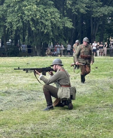 Dwaj żołnierze w historycznych mundurach i hełmach stoją na trawiastej polanie. Jeden klęczy i celuje ciężkim karabinem maszynowym w lewo, drugi idzie z karabinem w ręku. W tle gęsty las i grupa widzów stojących za taśmą.