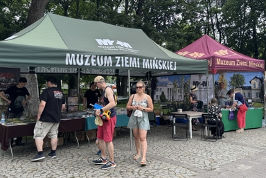 Dwa promocyjne namioty — zielony na pierwszym planie i bordowy z prawej. Namioty stoją na zewnątrz w cieniu drzew w ciągu dnia. Pod namiotami rozstawione są stoły z materiałami i ekspozycjami. Kilka osób rozmawia i ogląda stoiska. Na pierwszym planie kobieta w letniej sukience i okularach przeciwsłonecznych idzie pieszo, obok mężczyzna trzyma zabawkową ciężarówkę.