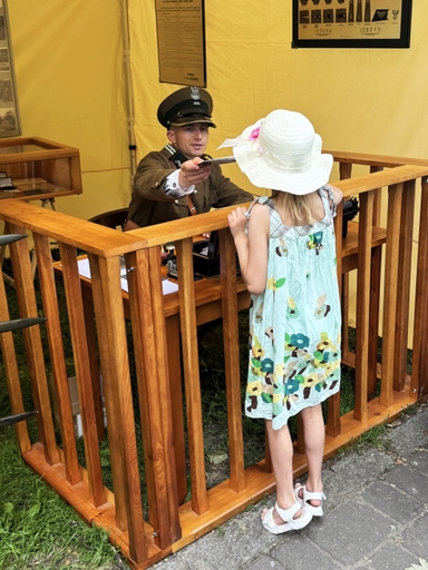 Mężczyzna w historycznym mundurze siedzi za drewnianą ladą i podaje dokument lub kartę. Przed ladą stoi dziewczynka w białym kapeluszu i jasnej sukience, żółte tło namiotu.