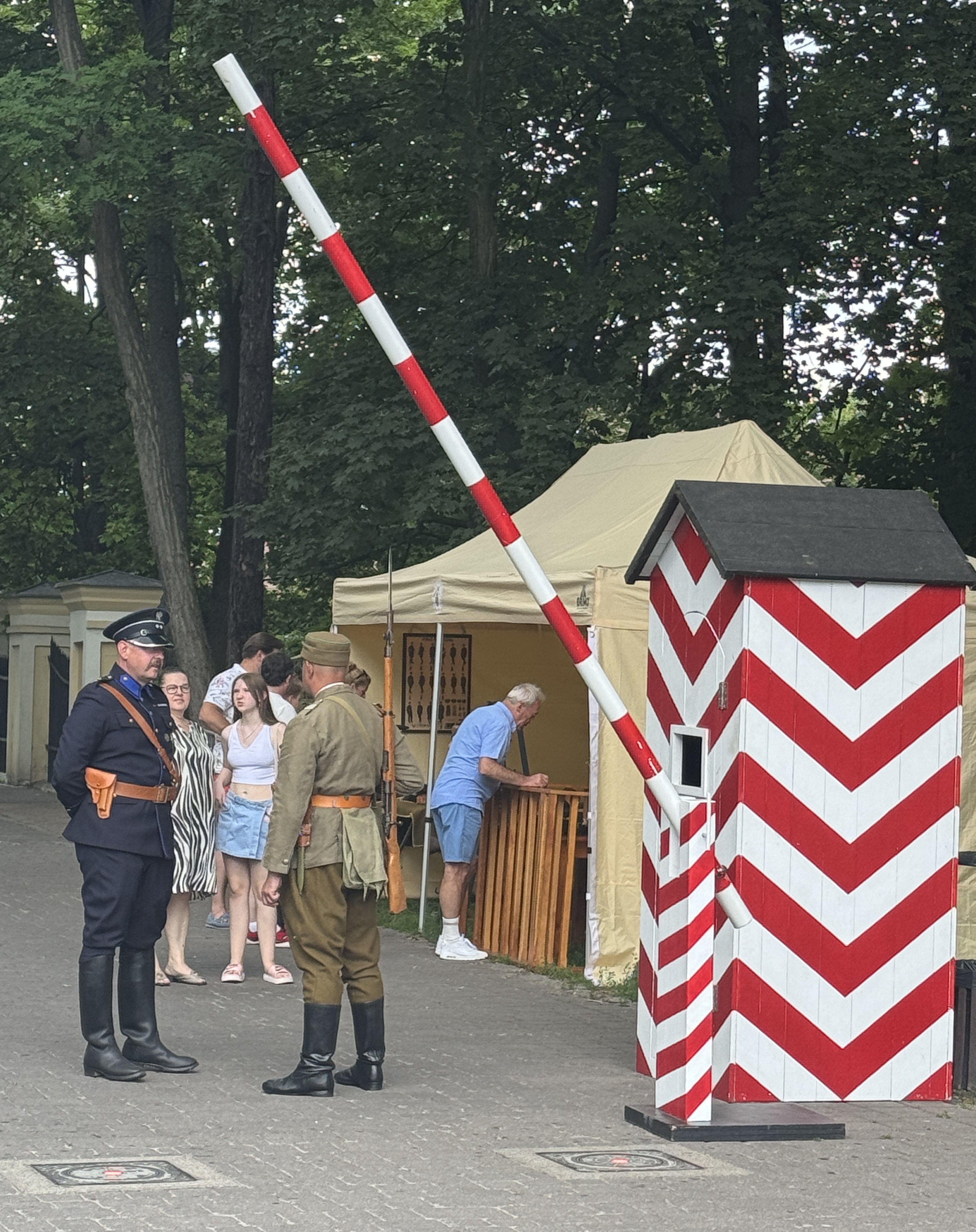 Czerwono-biały zapora wyciąga się ukośnie przez kadr. Po prawej stoi biało-czerwony wartowniczy budka z jaskrawym wzorem zygzaków. Na chodniku dwóch mężczyzn w historycznych mundurach rozmawia, za nimi stoi beżowy namiot i kilka osób pod drzewami.