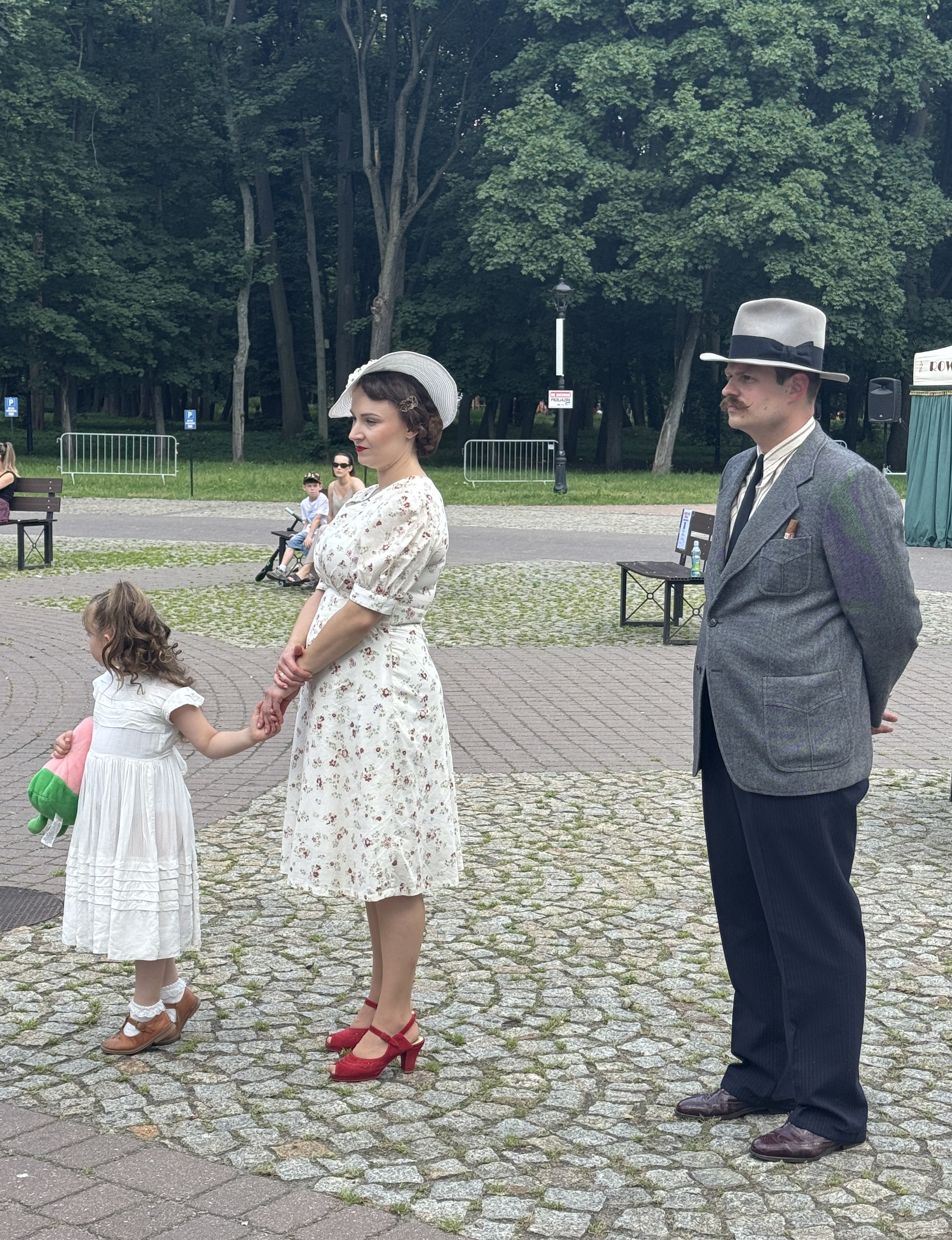 Kobieta w jasnej sukience w kwiaty i białym kapeluszu stoi trzymając za rękę małą dziewczynkę w białej sukience z pluszakiem. Mężczyzna w szarym garniturze, kapeluszu i wąsach stoi obok z rękami za plecami. Tło to park z gęstymi drzewami, chodnikiem z kostki i kilkoma ławkami.