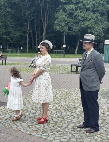 Kobieta w jasnej sukience w kwiaty i białym kapeluszu stoi trzymając za rękę małą dziewczynkę w białej sukience z pluszakiem. Mężczyzna w szarym garniturze, kapeluszu i wąsach stoi obok z rękami za plecami. Tło to park z gęstymi drzewami, chodnikiem z kostki i kilkoma ławkami.