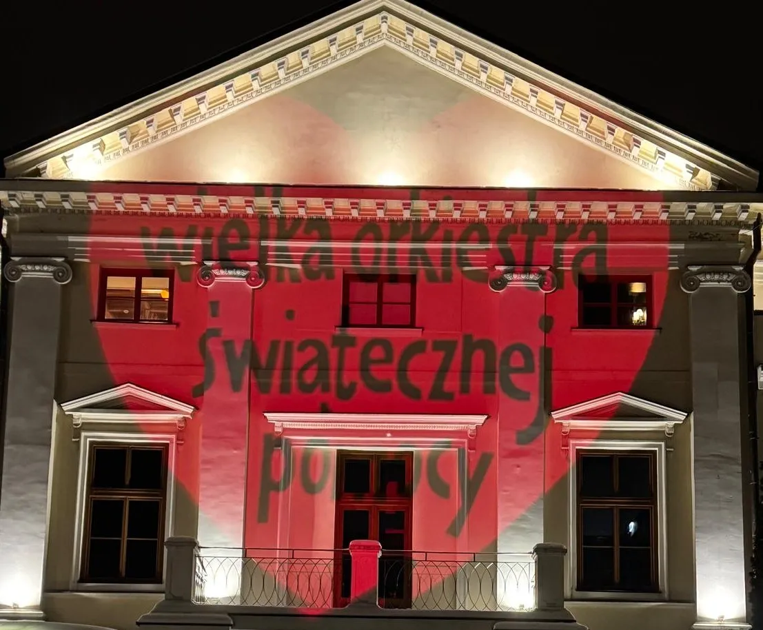 Budynek oświetlony nocą, na fasadzie biały klasyczny fronton i okna, z czerwonym sercem wyświetlonym na ścianie oraz czarnym napisem „wielka orkiestra świątecznej pomocy”.
