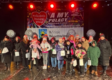 Grupa około 18 osób stoi na scenie na zewnątrz nocą, ubrana w zimowe ubrania i czapki, niektórzy trzymają białe torebki i bukiet kwiatów. W tle wisi kolorowy baner z logo Wielkiej Orkiestry Świątecznej Pomocy i napisem „Mińsk Mazowiecki”. Scenę oświetlają czerwone światła.