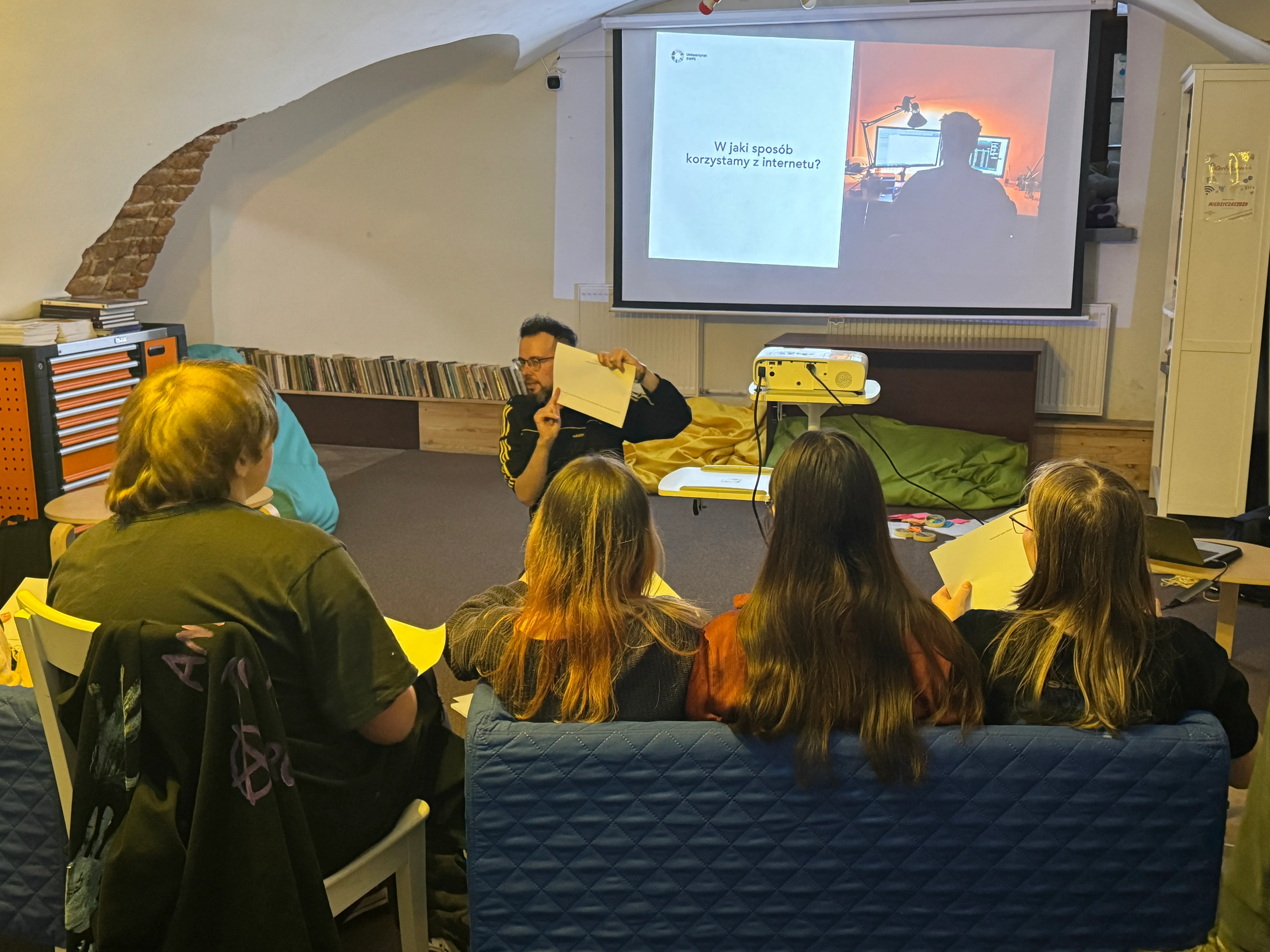 Cztery osoby siedzą tyłem na kanapie i krześle i oglądają prowadzącego. Prowadzący klęczy przed nimi i trzyma kartkę. Przed grupą stoi rzutnik i duży ekran z wyświetloną prezentacją. Pomieszczenie jest wewnątrz z niskim sklepieniem, półką z płytami i skrzynią narzędzi po lewej stronie.