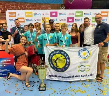 Grupa młodych zawodników z medalami i trenerami pozująca z pucharem i flagą Mińsko-Mazowieckiego Klubu Siatkarskiego na hali sportowej.