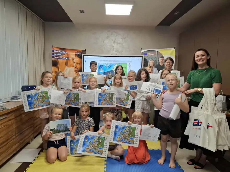 Grupa dzieci wraz z dorosłą osobą trzymająca kolorowe mapy Europy i flagi Unii Europejskiej w pomieszczeniu z biurkiem i tablicą interaktywną.