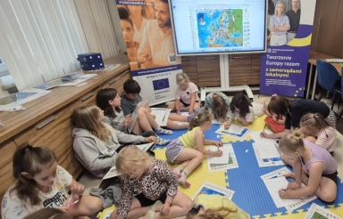 Dzieci siedzące i leżące na kolorowych matach rysują oraz wypełniają arkusze papieru w sali z mapą Europy wyświetloną na ekranie.