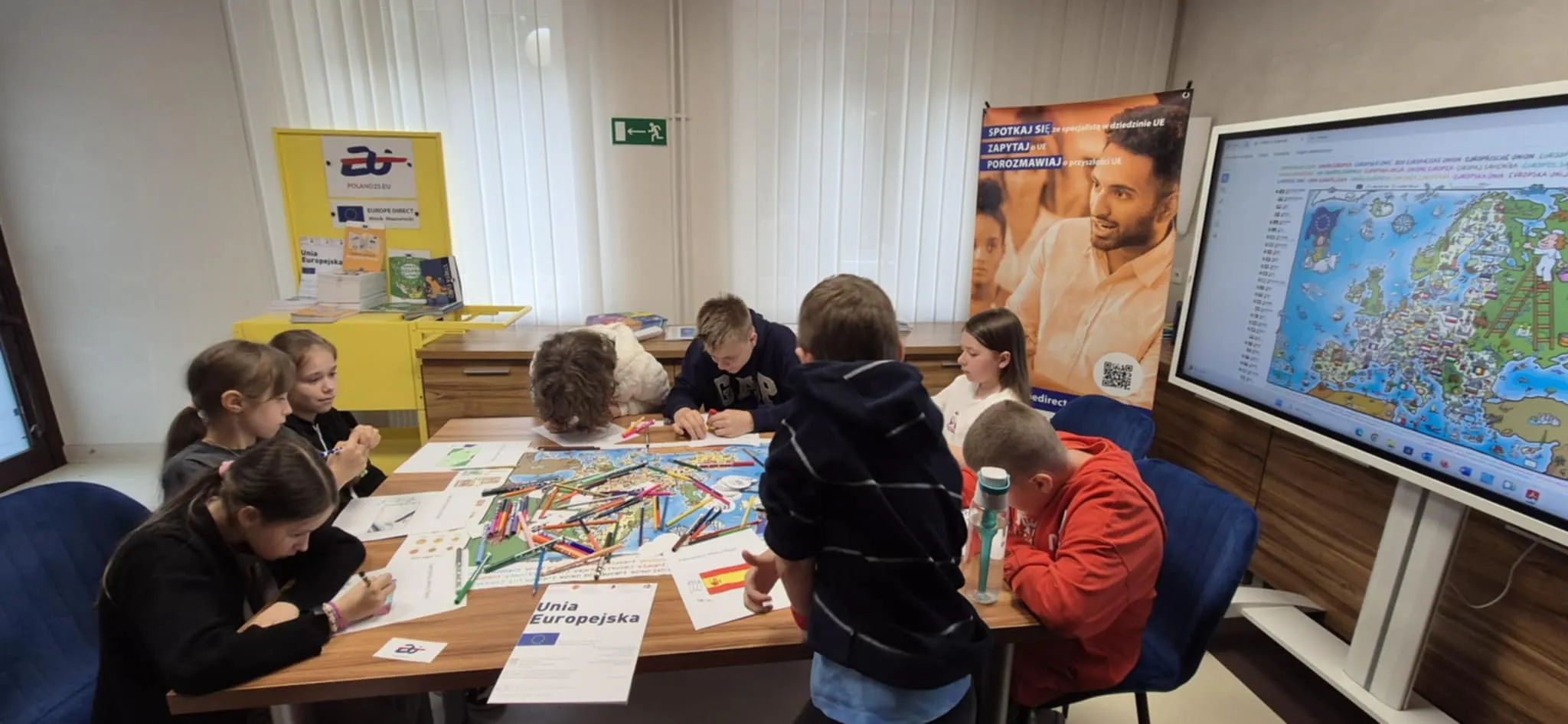 Grupa dzieci siedzi i stoi przy stole, kolorując mapę Europy oraz wypełniając arkusze papieru.