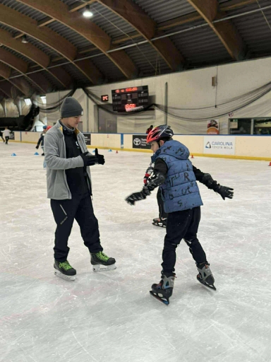 Dwóch mężczyzn jeździ na łyżwach po lodowisku krytym. Jeden ma szarą czapkę i kurtkę, drugi czerwoną kask i niebieską kamizelkę. W tle widać tarczę z zegarem i tablice reklamowe.