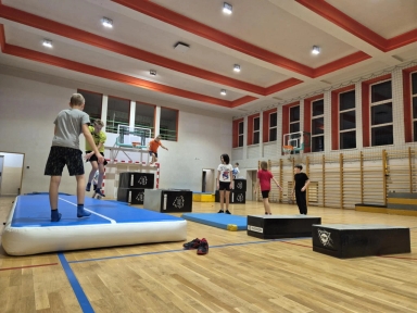 Wewnątrz hali sportowej dzieci ćwiczą na materacach i skrzyniach gimnastycznych rozstawionych na drewnianej podłodze. W tle widać kosze do koszykówki oraz ściankę do wspinaczki z drabinkami. Pomieszczenie jest jasne, z białymi ścianami i czerwonymi elementami na suficie oraz wokół okien.