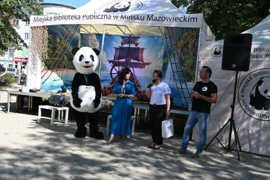 Stoiska plenerowe Miejskiej Biblioteki Publicznej w Mińsku Mazowieckim z ludźmi, postacią w kostiumie pandy oraz dużym muralem z żaglowcem w tle, na zewnątrz w dzień.
