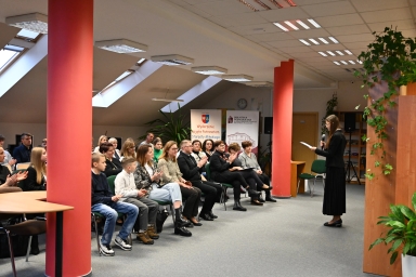 Grupa około dwudziestu osób siedzi w rzędach na zielonych krzesłach w jasnym pomieszczeniu z dużymi oknami dachowymi. Po prawej stronie stoi młoda kobieta w czarnym stroju, trzyma mikrofon i czyta z kartki. W tle widoczne są dwa czerwone filary, plakaty oraz rośliny doniczkowe.