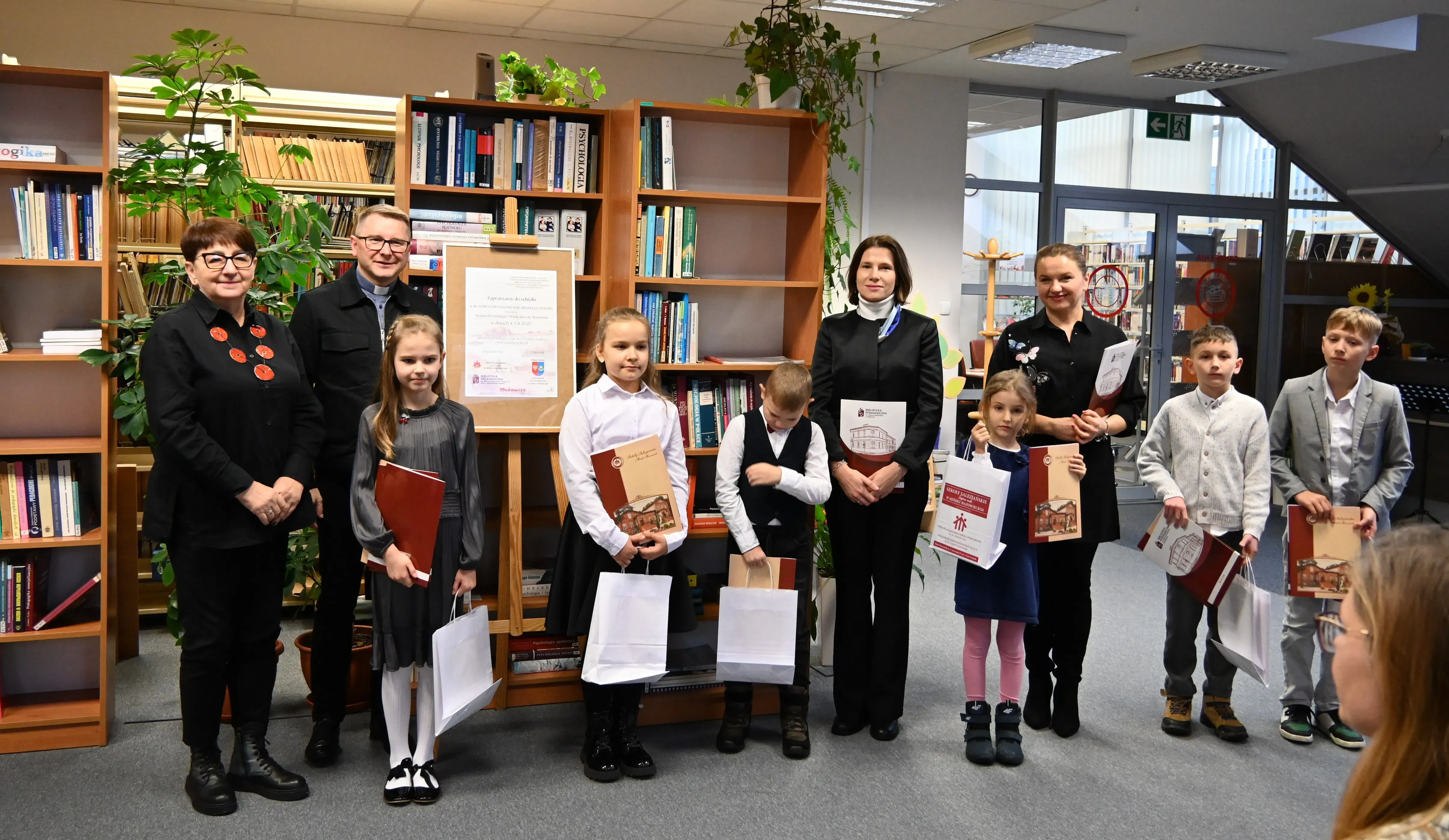 Grupa sześciu dzieci i czterech dorosłych stoi w bibliotece przed regałami z książkami. Dzieci trzymają białe torby i teczki. W tle jest plansza z napisem oraz rośliny doniczkowe. Pomieszczenie jest dobrze oświetlone sztucznym światłem.