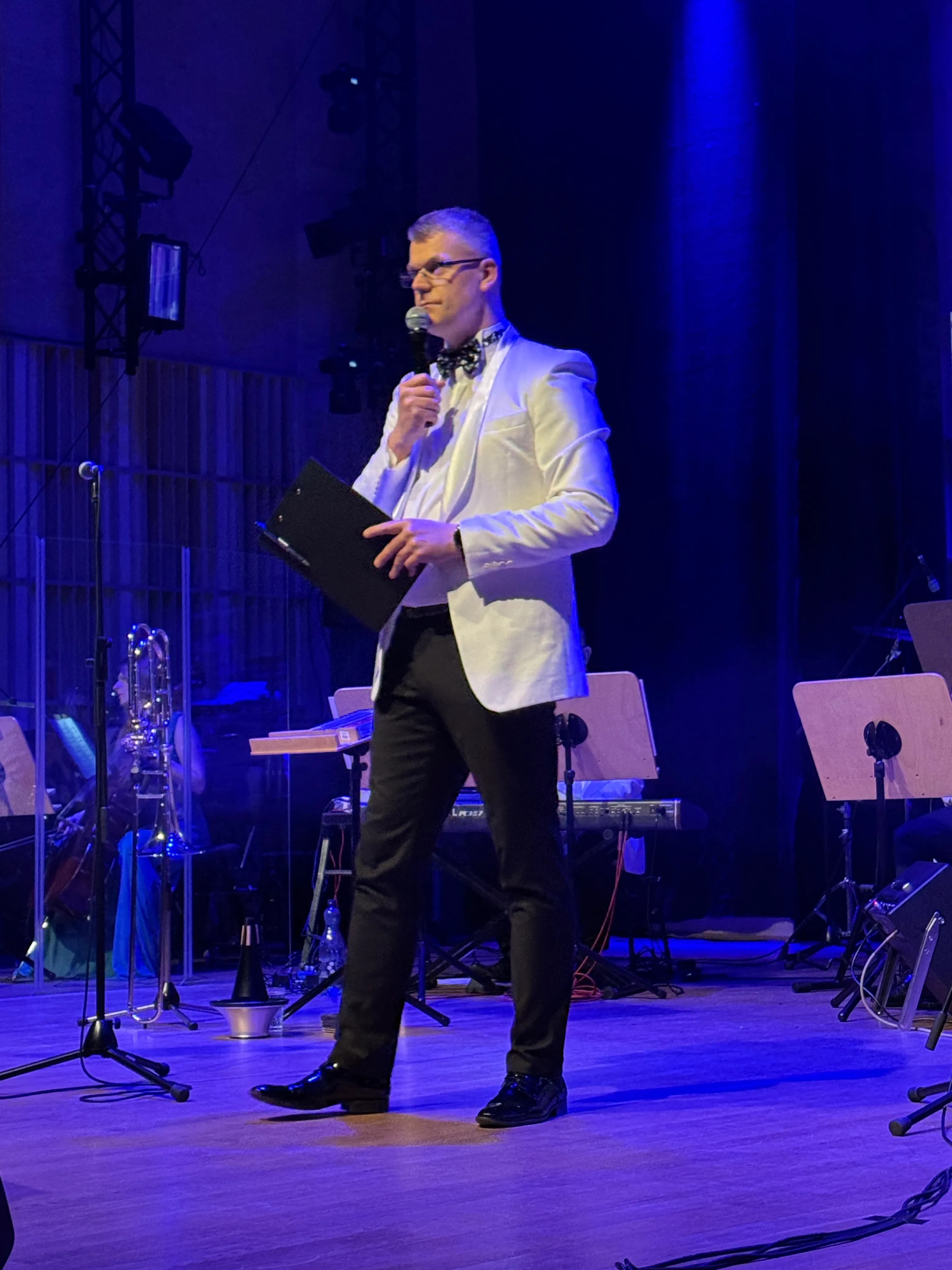 Mężczyzna w białej marynarce i czarnych spodniach stoi na scenie. Trzyma mikrofon przy ustach i czarną teczkę. Scena jest oświetlona niebieskim światłem, w tle stoją stojaki z nutami i instrumenty orkiestry.
