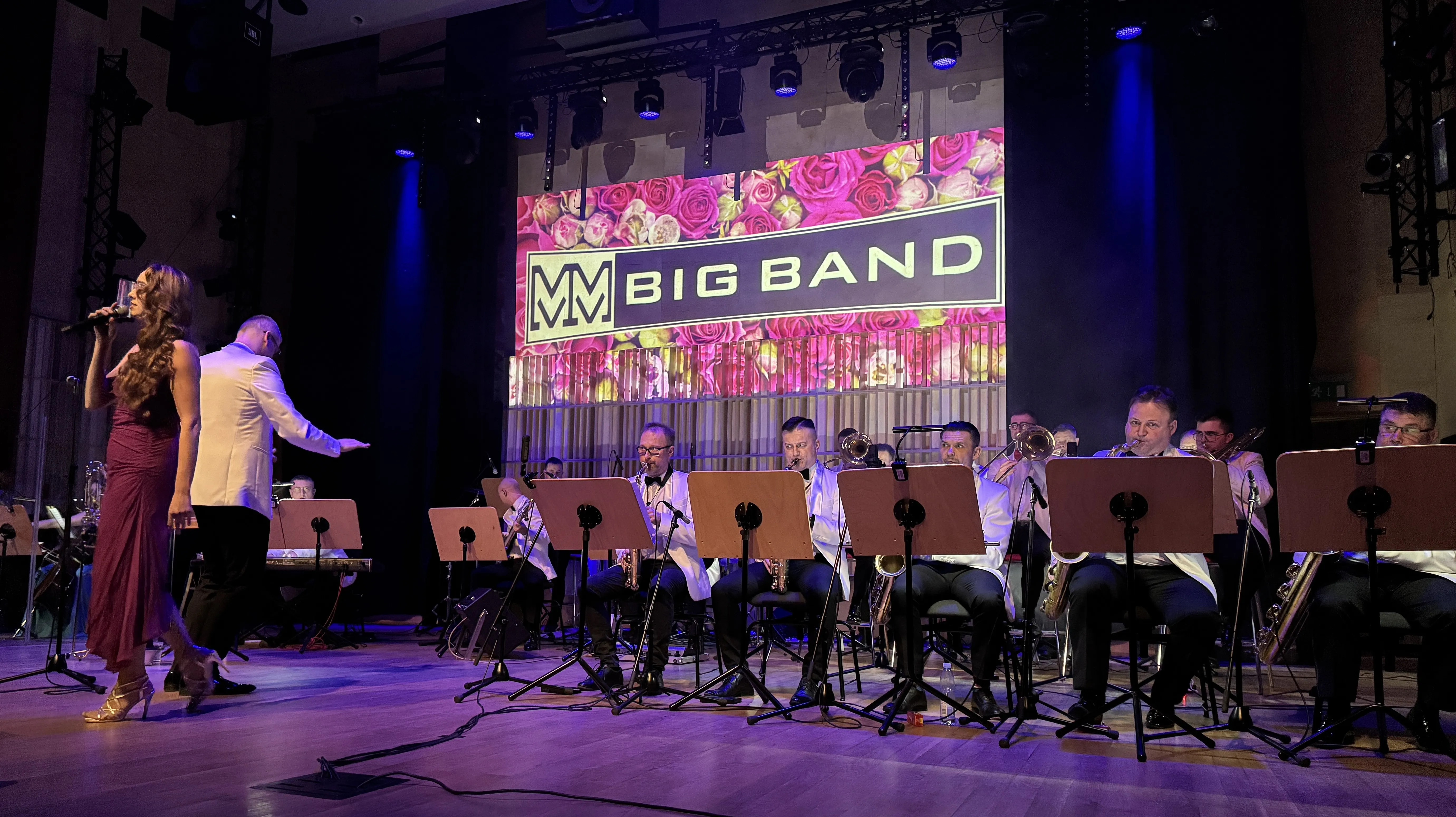 Kobieta w długiej sukni śpiewa przy mikrofonie po lewej stronie. Mężczyzna w białej marynarce dyryguje stojąc obok niej. Grupa muzyków w białych marynarkach siedzi w rzędzie na scenie i gra na dętych instrumentach. Za nimi wisi duży prostokątny banner z różowym, kwiatowym motywem i prostą grafiką logo. Scena ma ciemne tło i fioletowe oświetlenie.