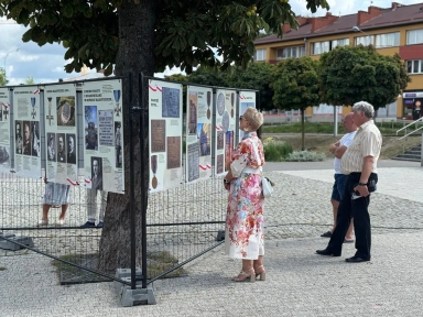 Na zewnątrz. Trzy osoby stoją przy planszach z wystawą historyczną umieszczoną wokół drzewa na placu z kostki brukowej, w tle drzewa i budynki mieszkalne.