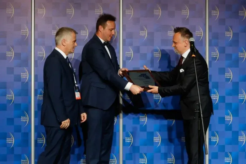 Trzech mężczyzn w garniturach na tle niebieskiej ścianki, jeden przekazuje płaską nagrodę drugiemu.