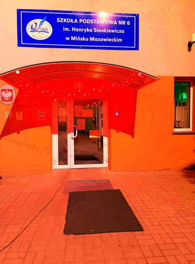 Wejście do budynku szkoły podstawowej z białymi drzwiami, nad którym znajduje się niebieska tablica z nazwą szkoły na pomarańczowym tle. Po prawej stronie okno, a przed wejściem położona jest czarna mata na czerwonej kostce brukowej.