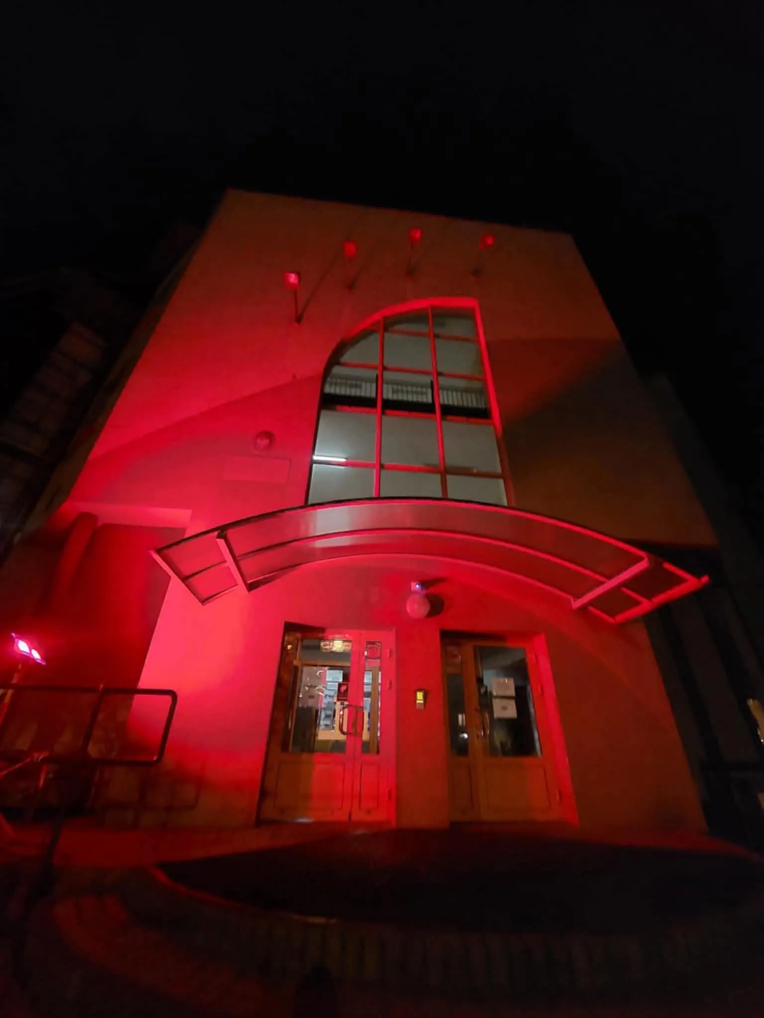 Budynek z dużym, półkolistym daszkiem nad wejściem, podświetlony czerwonym światłem w nocy, z dwoma drzwiami i dużym oknem nad nimi.