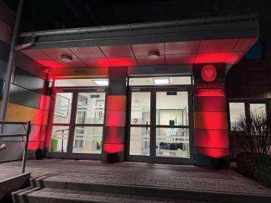 Wejście do budynku w nocy, oświetlone na czerwono wokół drzwi. Przed budynkiem schody i poręcz, po prawej stronie krzewy. Wnętrze widoczne przez przeszklone drzwi dobrze oświetlone.