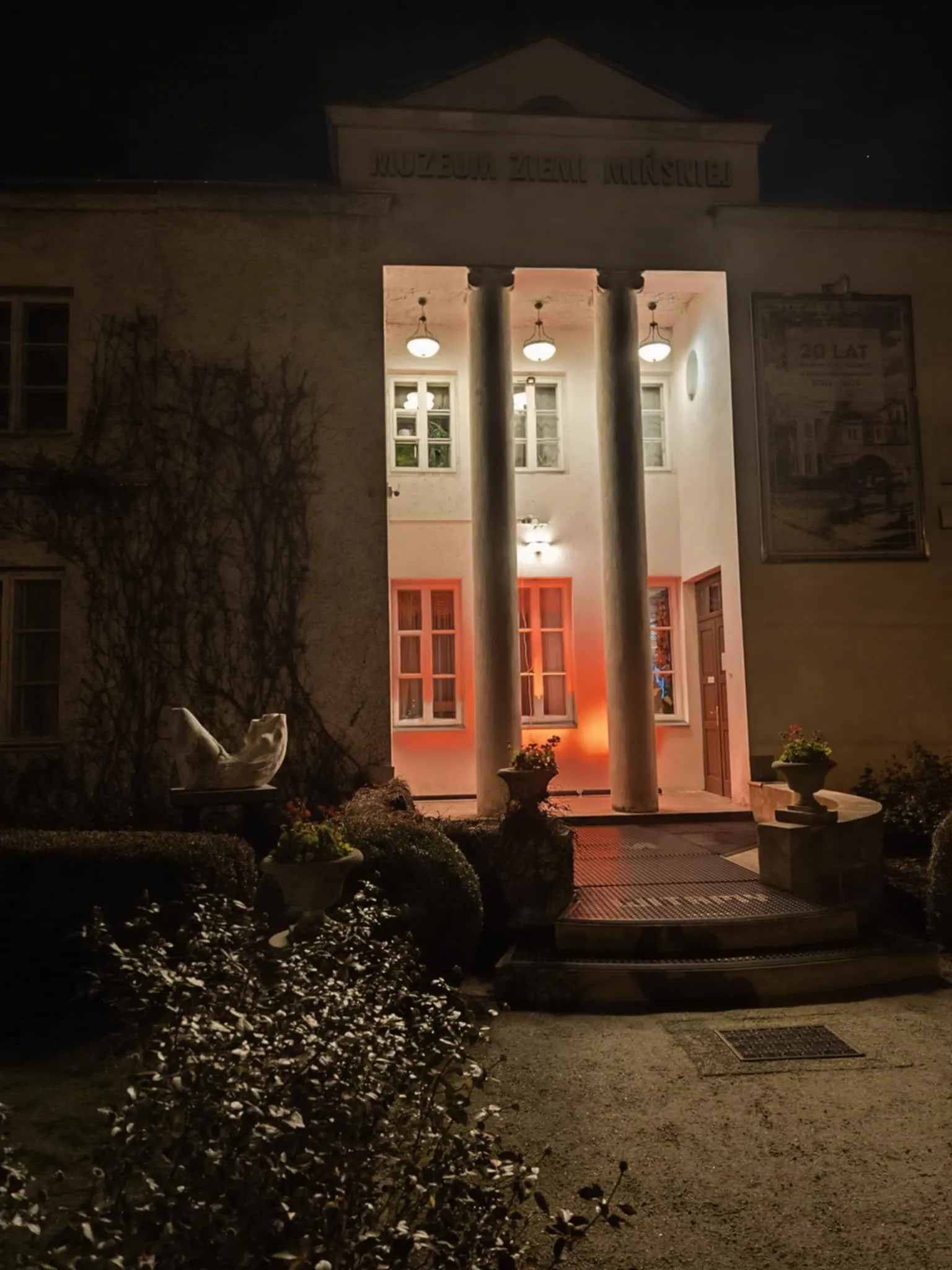 Wejście do budynku muzeum oświetlone nocą, dwie kolumny przy wejściu, po bokach donice z roślinami, przy ścianie rzeźba, na elewacji pnącza.