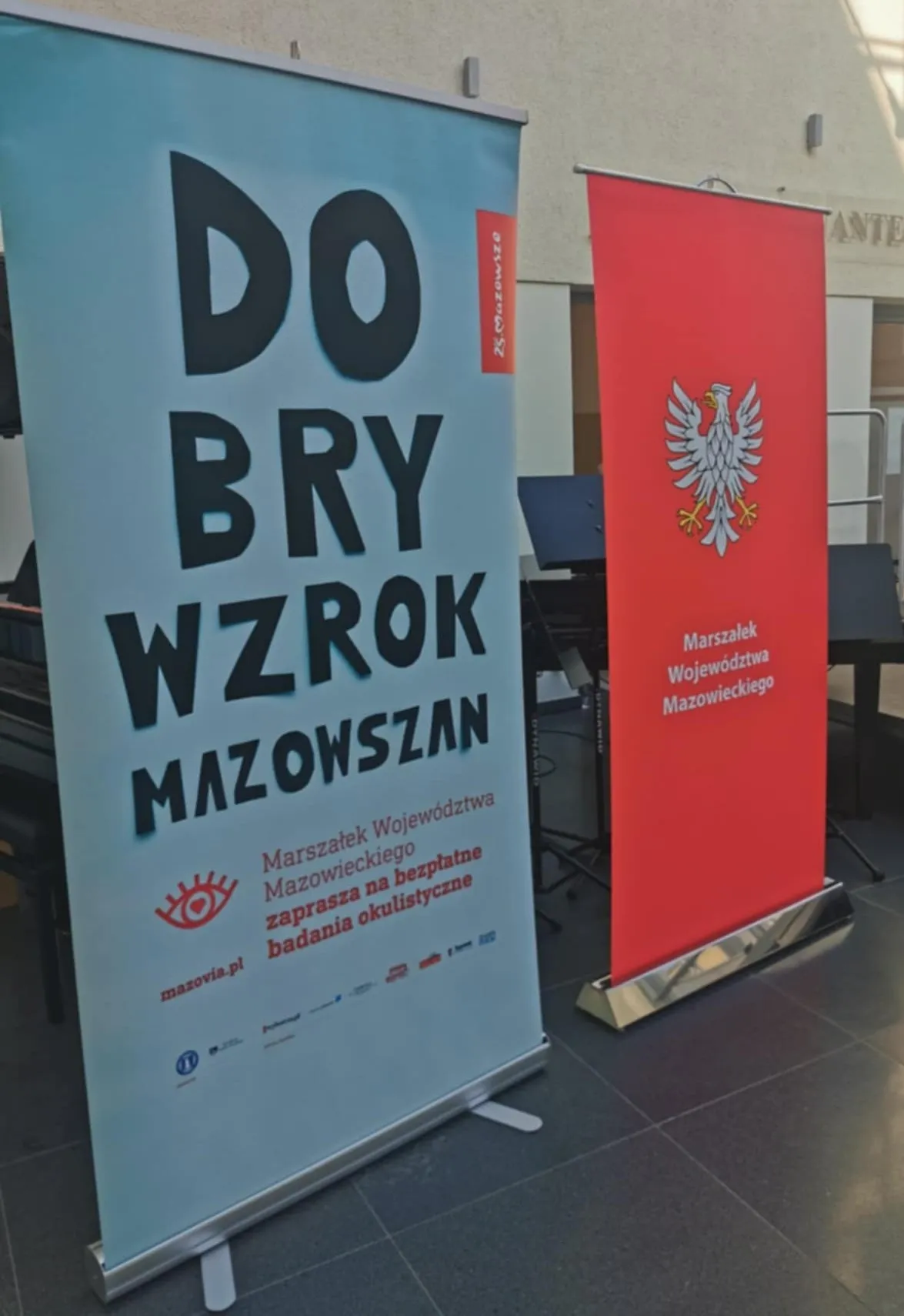 Dwa stojące roll-upy: niebieski z napisem zachęcającym do bezpłatnych badań okulistycznych i czerwony z godłem Polski oraz napisem Marszałek Województwa Mazowieckiego.