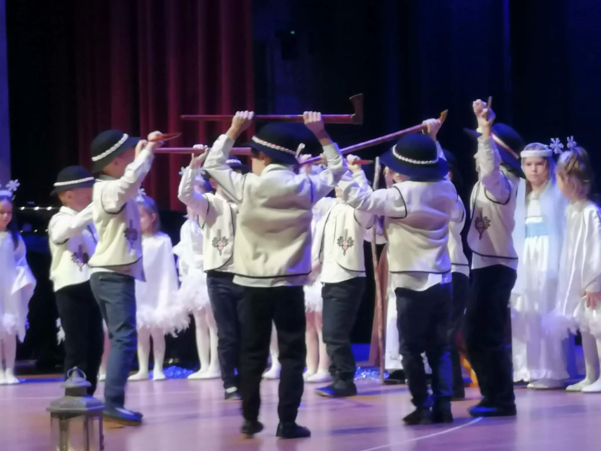 Grupa dzieci na scenie podczas występu. Chłopcy w tradycyjnych strojach ludowych z czarnymi kapeluszami trzymają długie kije nad głowami. W tle dziewczynki ubrane na biało, część z nich ma ozdoby na głowach. Scena ma drewnianą podłogę, za dzieci stoi czerwony zasłon i ciemne tło.