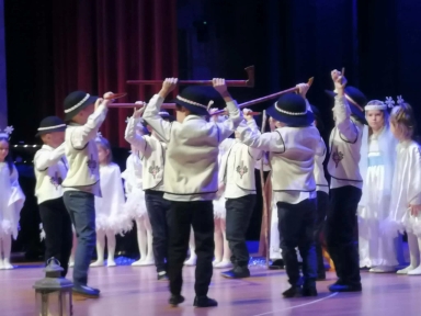 Grupa dzieci na scenie podczas występu. Chłopcy w tradycyjnych strojach ludowych z czarnymi kapeluszami trzymają długie kije nad głowami. W tle dziewczynki ubrane na biało, część z nich ma ozdoby na głowach. Scena ma drewnianą podłogę, za dzieci stoi czerwony zasłon i ciemne tło.