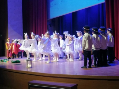 Na scenie występuje grupa dzieci w kolorowych strojach. Po lewej stronie dzieci ubrane na czerwono i żółto, w centrum dziewczynki w białych sukienkach z dekoracjami na głowach, po prawej chłopcy w tradycyjnych strojach z kapeluszami. Scena oświetlona jest niebieskim światłem, tło ciemne z czerwonymi zasłonami.