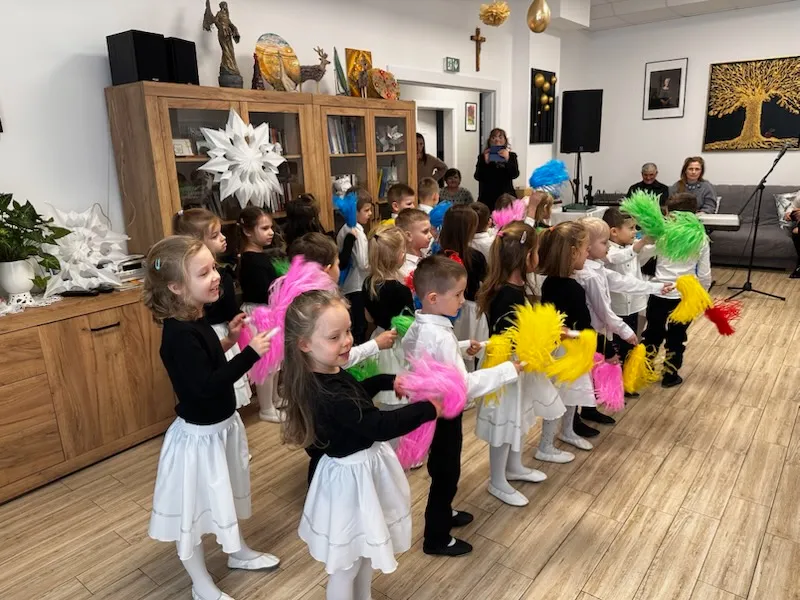 Mężczyzna siedzi po prawej i gra na gitarze przy mikrofonie i statywie z nutami. Grupa około dziesięciu dzieci siedzi na podłodze po lewej i słucha. Kilka dorosłych siedzi na krzesłach na pierwszym planie. Jasne wnętrze dzienne z białymi zasłonami, złotymi i białymi dekoracjami wiszącymi oraz dużą drewnianą szafką z książkami i ozdobami z tyłu.