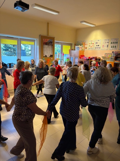Grupa kilkunastu dorosłych osób trzyma się za ręce i tańczy w kole w jasnej sali z beżowymi ścianami. W tle widać okna z żółtymi roletami oraz półki z zabawkami i kolorowymi plakatami na ścianie.