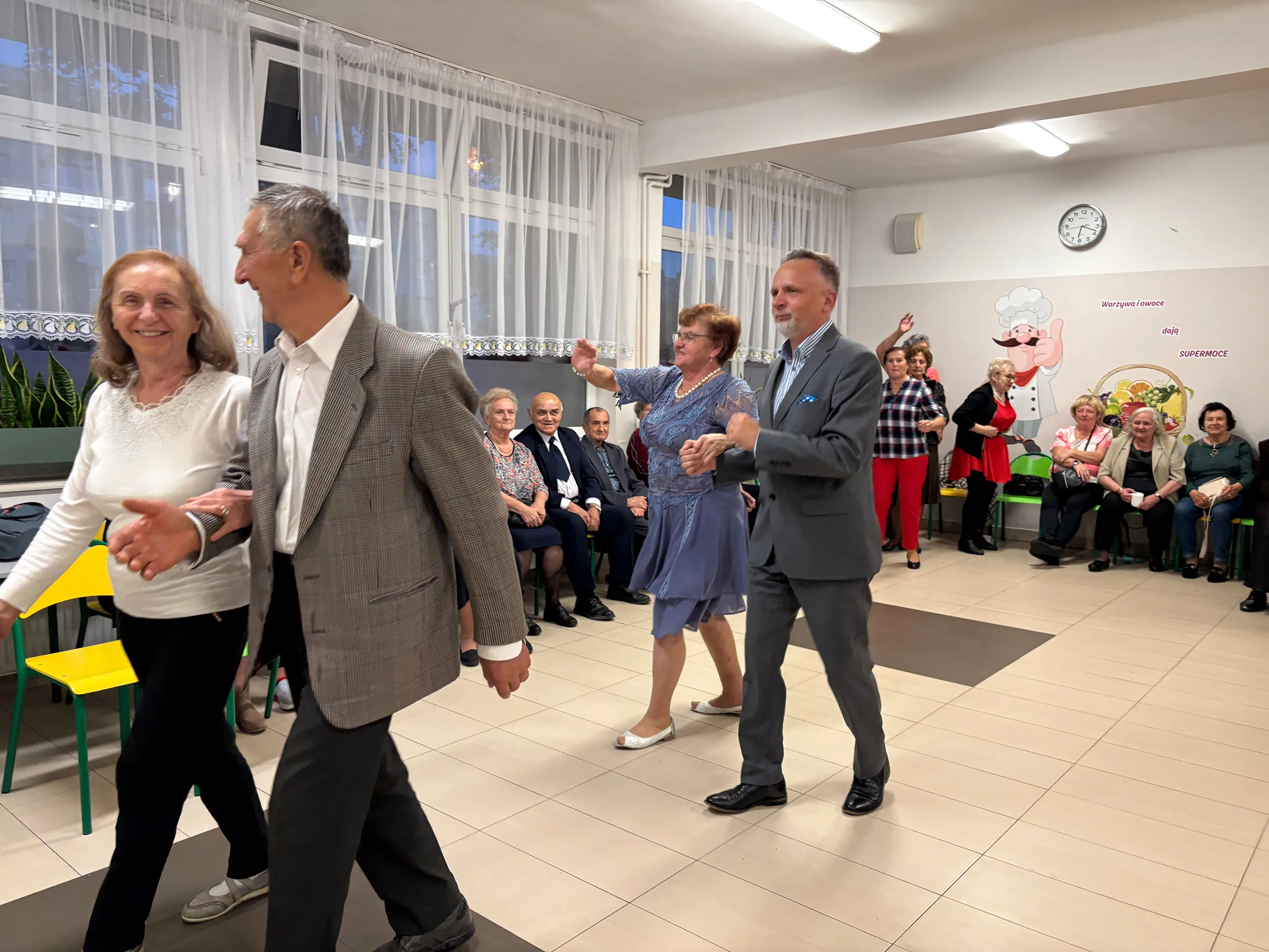 W sali z jasnymi ścianami i dużymi oknami tańczą dwie pary starszych osób, jedna para idzie na przód po lewej, druga – po prawej. W tle kilkanaście osób siedzi lub stoi przy ścianie. Na ścianie znajduje się zegar i kolorowa grafika z postacią kucharza oraz koszem owoców.