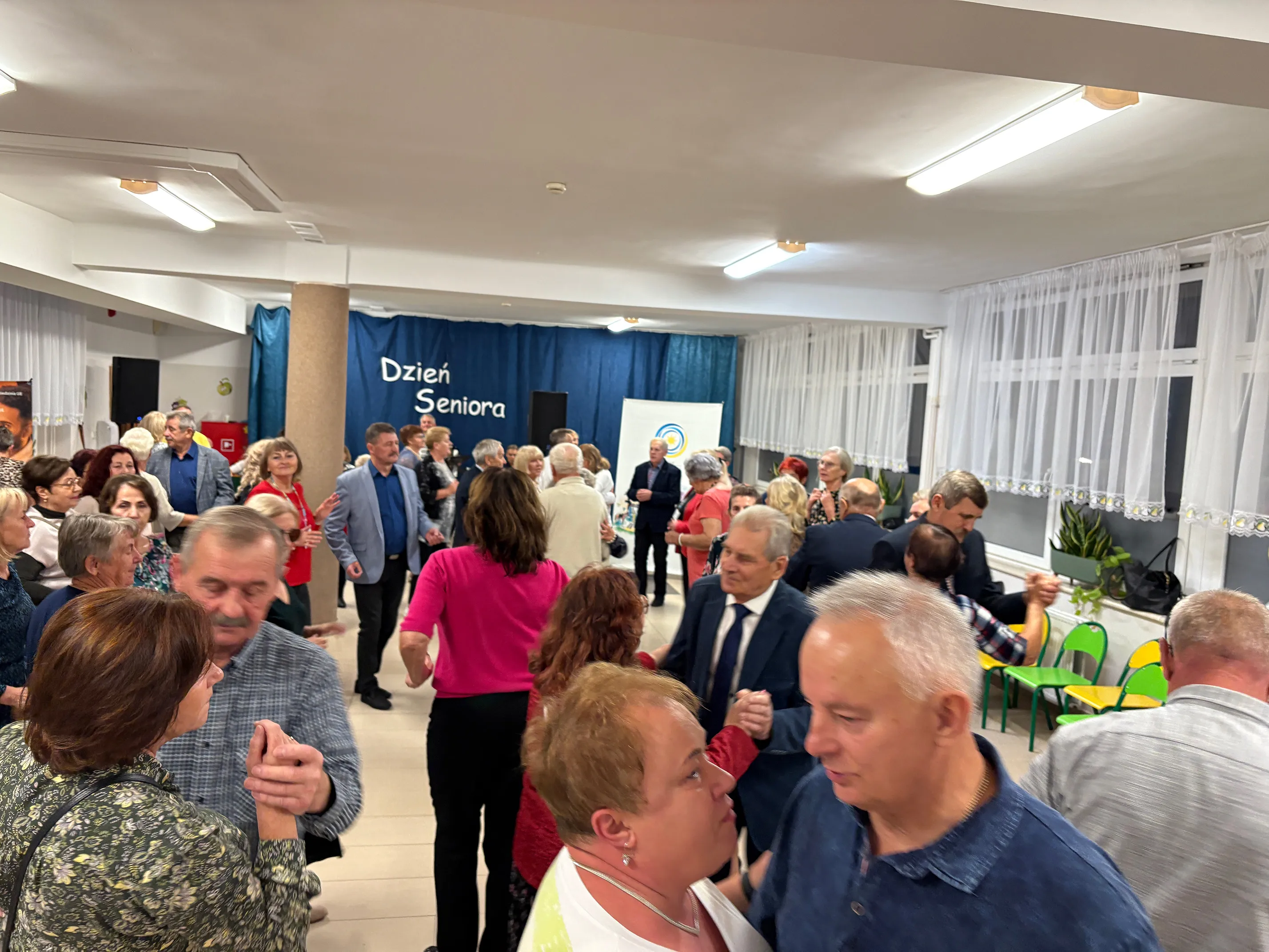 Grupa starszych osób tańczy w jasnym, dużym pomieszczeniu z białymi ścianami i oknami udekorowanymi firankami. W tle widoczna granatowa kurtyna z napisem „Dzień Seniora”. Niektóre osoby są w parach, na środku sali. Po prawej stronie widać rząd zielono-żółtych krzeseł i rośliny na parapecie.