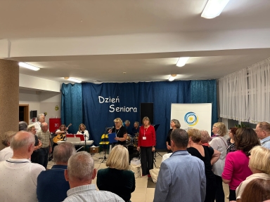 Grupa starszych osób stoi w pomieszczeniu z jasnym sufitem i oświetleniem. Na środku stoi kobieta z mikrofonem, obok niej inna kobieta w czerwonej bluzce. W tle niebieska zasłona z napisem "Dzień Seniora" oraz stojak z logo. Z lewej strony osoby grają na gitarach i keyboardzie. Po prawej stronie okno z białą firaną.