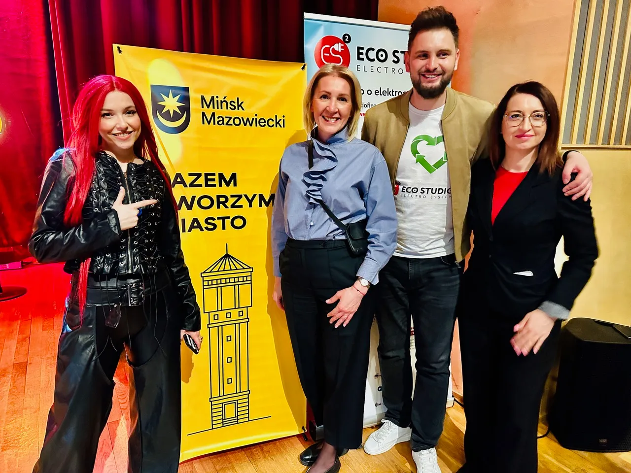 Cztery osoby stoją w sali z drewnianą podłogą przed żółtym banerem z herbem miasta i rysunkiem wieży. Po lewej kobieta z długimi czerwonymi włosami i czarną kurtką wskazuje palcem. Druga od lewej kobieta w jasnoniebieskiej bluzce stoi z torebką przez ramię. Mężczyzna w białej koszulce z zielonym symbolem trzyma rękę na ramieniu kobiety po prawej. Po prawej kobieta w czarnym garniturze i okularach stoi z ręką u boku.