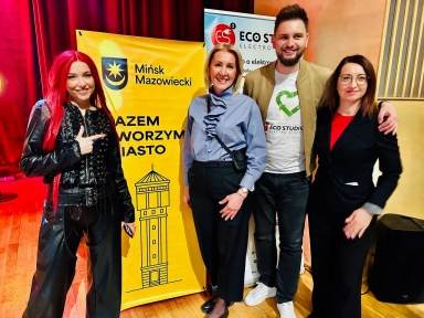 Cztery osoby stoją w sali z drewnianą podłogą przed żółtym banerem z herbem miasta i rysunkiem wieży. Po lewej kobieta z długimi czerwonymi włosami i czarną kurtką wskazuje palcem. Druga od lewej kobieta w jasnoniebieskiej bluzce stoi z torebką przez ramię. Mężczyzna w białej koszulce z zielonym symbolem trzyma rękę na ramieniu kobiety po prawej. Po prawej kobieta w czarnym garniturze i okularach stoi z ręką u boku.